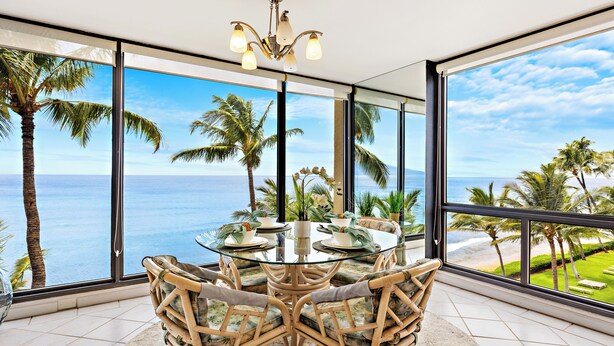 Ein Essbereich mit einem Glastisch und Korbstühlen, große Fenster mit Blick auf das Meer und Palmen.