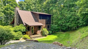 A log cabin with a stone chimney, surrounded by lush greenery and a well-maintained lawn. - Bearly Behaving | 3BR Cabin w/ Hot Tub, Pool Table, Fire Pit (Todd)