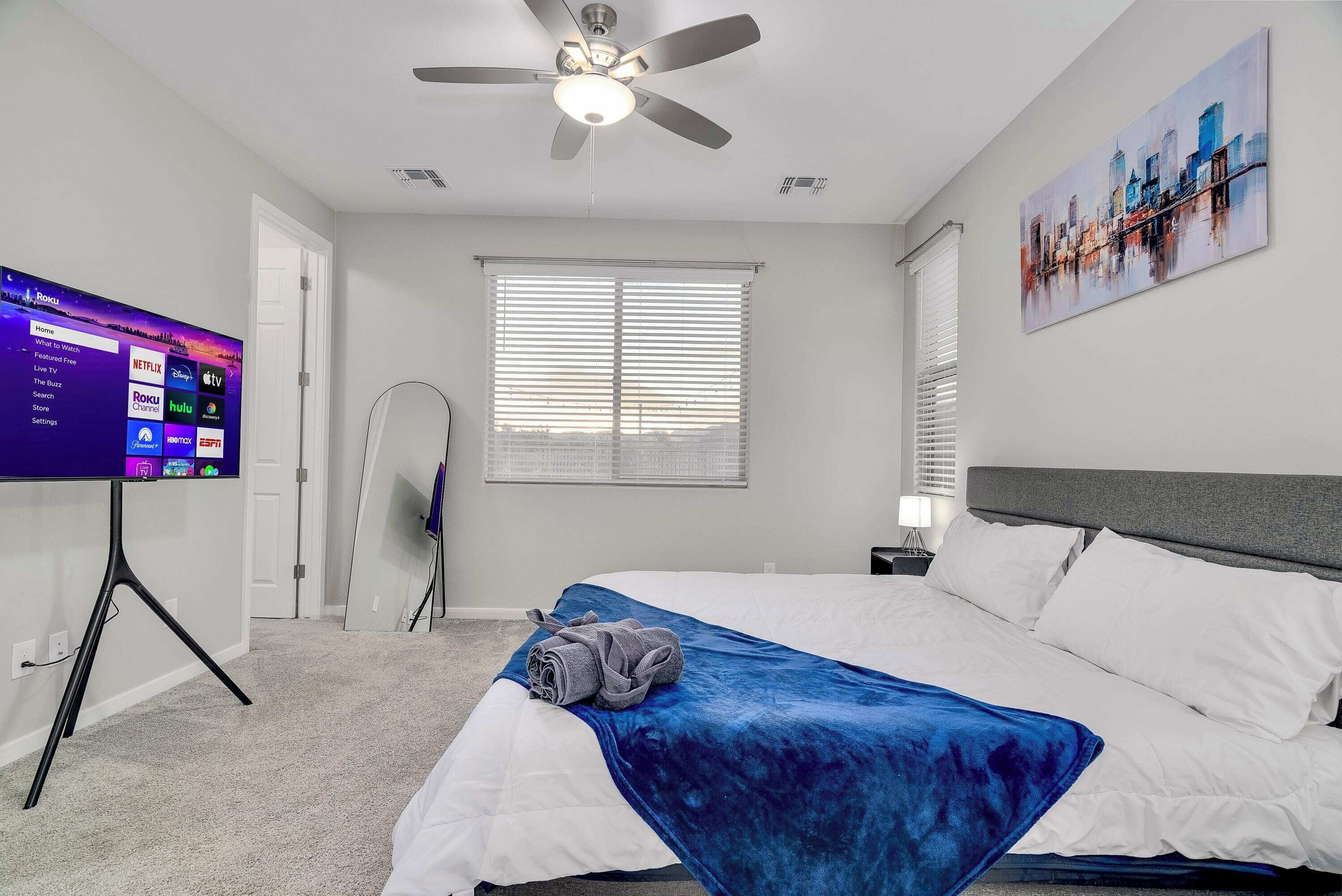 A hotel room with a bed, a ceiling fan, a TV displaying streaming services, a window with blinds, and a cityscape painting on the wall.