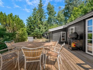 A wooden deck with a round table and chairs, a barbecue grill, and a view of a forested area. - "Synnöve" - 350m from the sea by Interhome (Væggerløse)