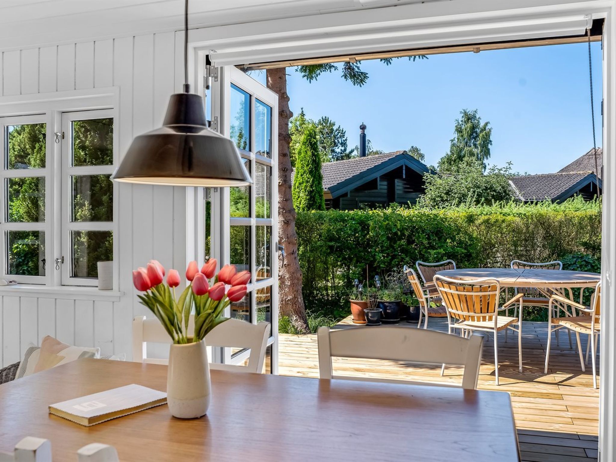 A wooden deck with a table and chairs, a hanging pendant light, and a view of a house and greenery.
