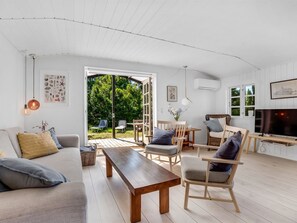 Amplia sala de estar con un banco de madera, un sofá y dos sillones. Hay un televisor sobre un mueble de madera, y un ventanal con vistas al verdor exterior.