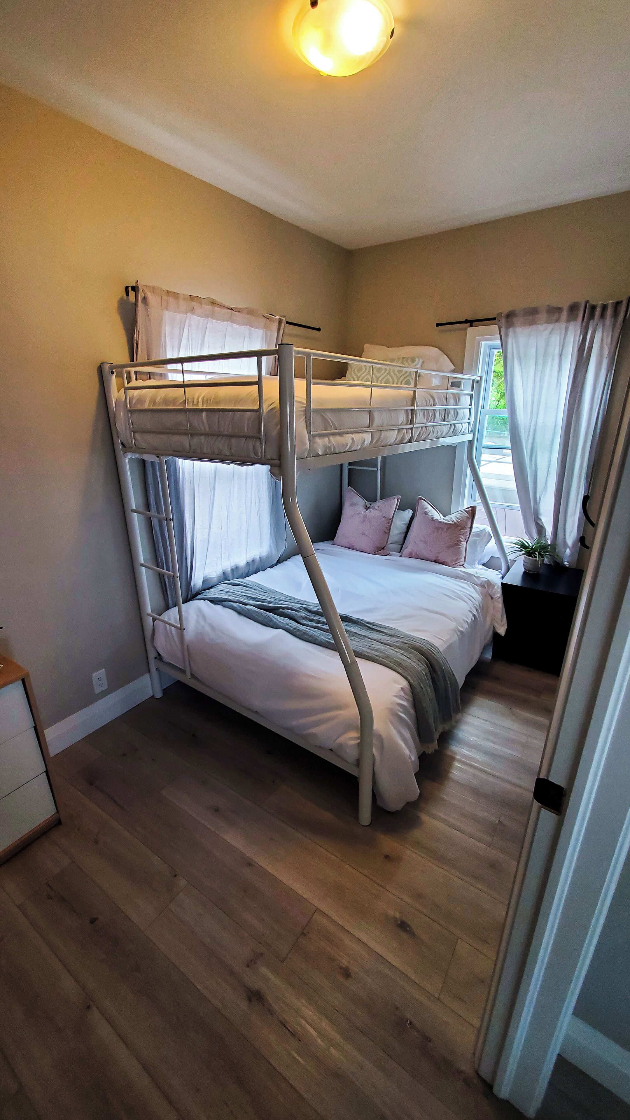 A bunk bed room with a wooden floor, a bedside table with a plant, and a window with curtains.