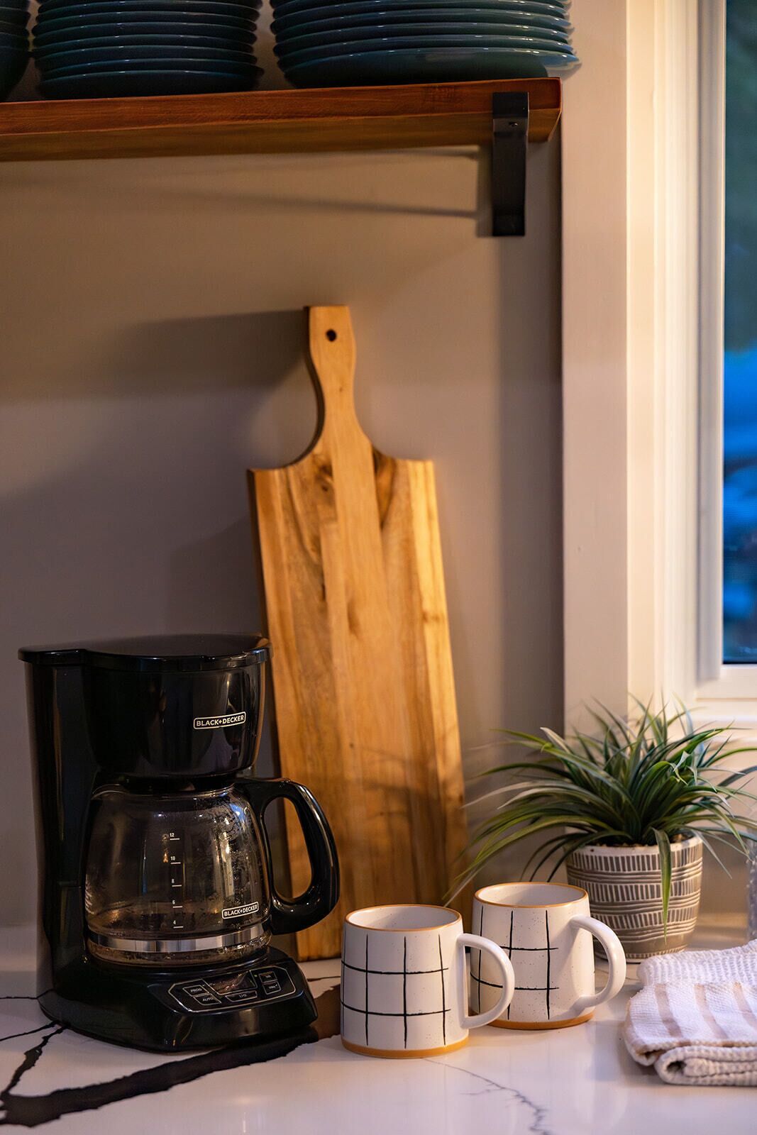 Une cuisine équipée d’une machine à café, de deux tasses, d’une planche à découper et d’une plante en pot.