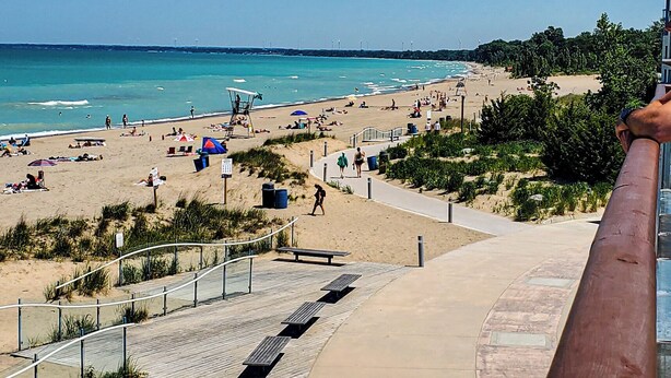 Una playa con un paseo marítimo de madera, gente disfrutando y un cielo azul despejado.
