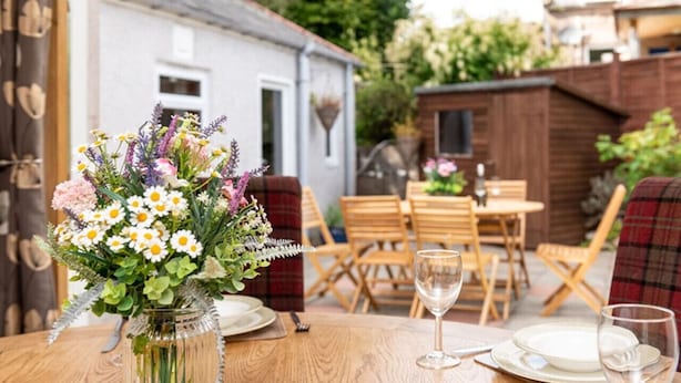 A wooden table set for dining with a vase of flowers, a wooden chair, and a dining area with a view of a house and garden. - Dormie Cottage, spacious bungalow with log fire! (Aberdeenshire)