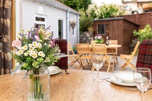 A wooden table set for dining with a vase of flowers, a wooden chair, and a dining area with a view of a house and garden. - Dormie Cottage, spacious bungalow with log fire! (Aberdeenshire)