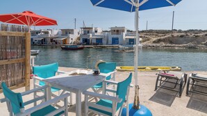 Een terras aan zee met witte tafels en blauwe stoelen, met uitzicht op een rustige haven met boten en witte gebouwen.