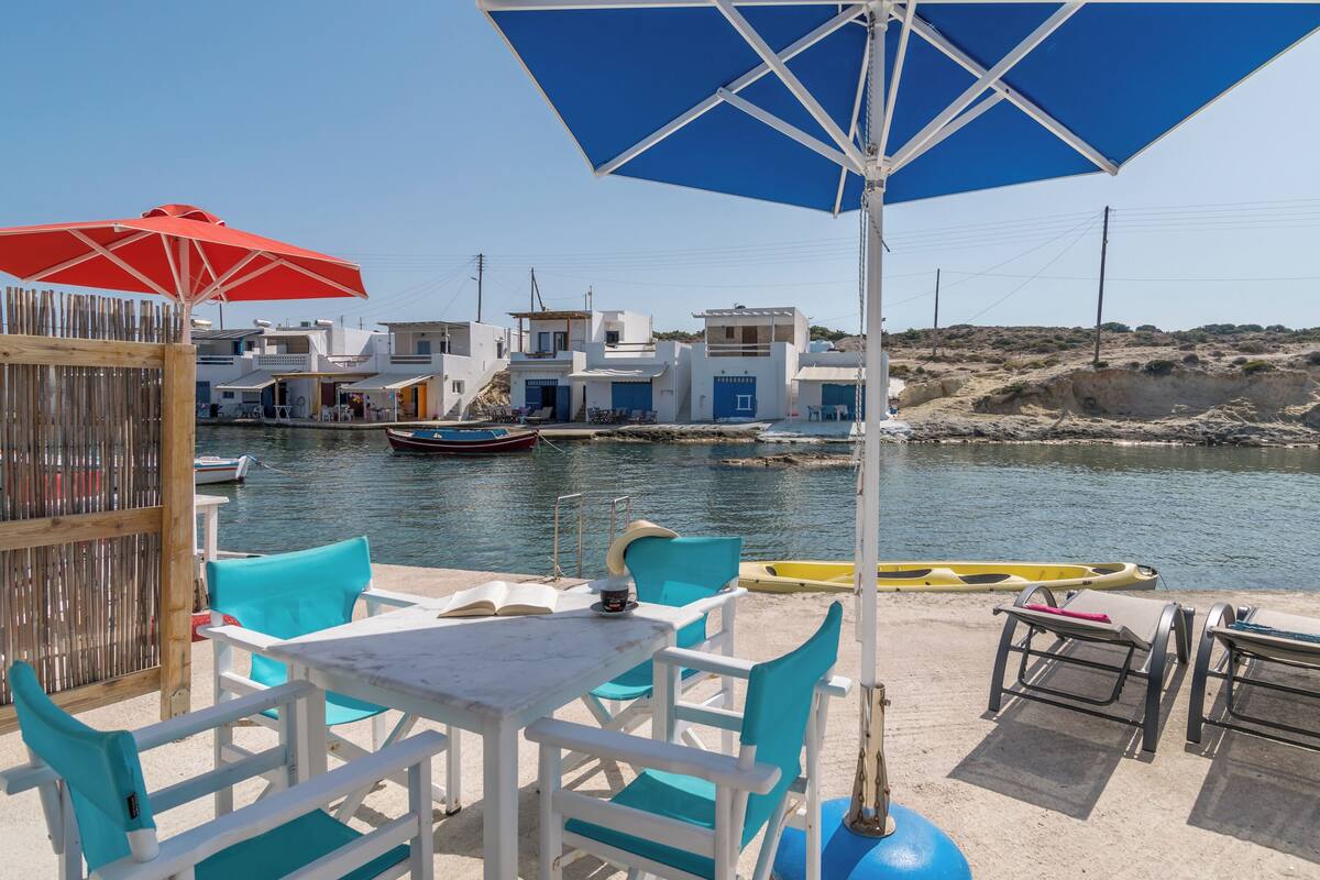 A seaside terrace with white tables and blue chairs, overlooking a calm harbor with boats and white buildings.