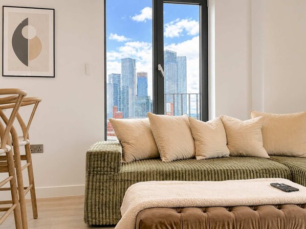 A modern bedroom with a green upholstered sofa bed, a wooden chair, and a city view through a large window.
