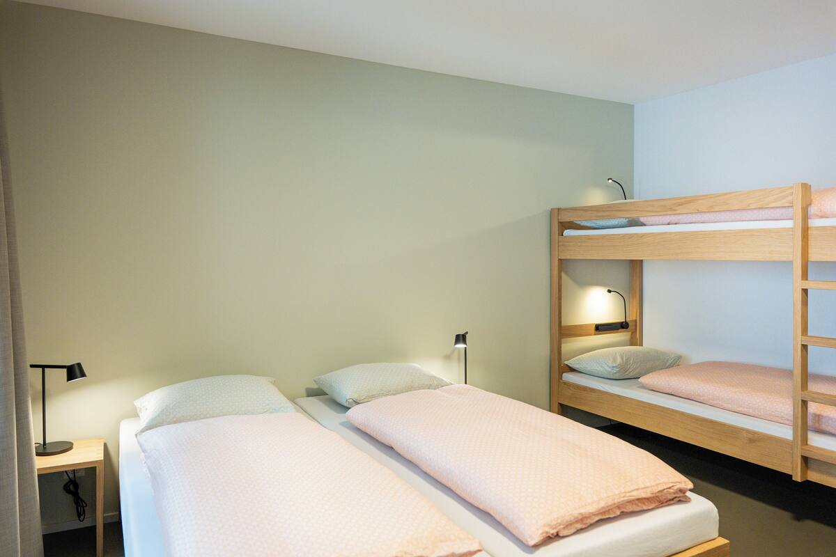 A room with two single beds and a bunk bed, each with pillows and blankets. There is a nightstand with a lamp and a power outlet.