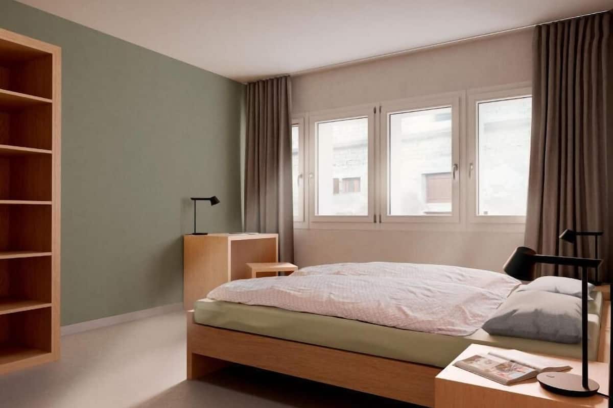 A bedroom with a bed, a bedside table, a lamp, a wooden shelf, and a window with curtains.