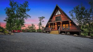 A wooden cabin with a triangular roof, a porch, and a gravel driveway.