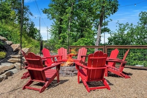 A fire pit with red Adirondack chairs arranged around it, set on a gravel surface with trees and a wooden fence in the background.
