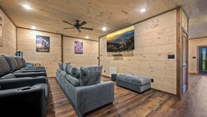 A room with a wooden ceiling, ceiling fan, and a large wall-mounted TV. There are two sofas and a small ottoman.