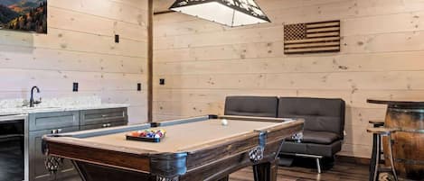 Una mesa de billar con acabados de madera, un sofá, una zona de bar y una pared de madera con una bandera.