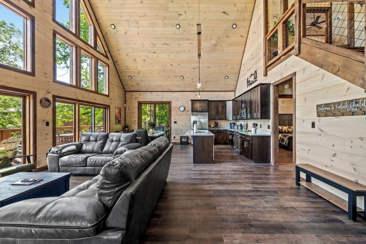 A spacious living area with a large leather sofa, wooden flooring, and a kitchen in the background.