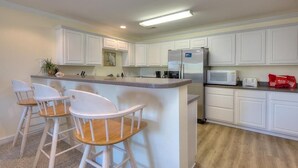 A kitchen with white cabinets, a stainless steel refrigerator, a microwave, and a bar area with three white bar stools. - Enjoy beach walks and family fun in this 2BR, 2BA Oak Island condo. (Oak Island)