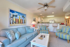 A living room with a sofa, armchairs, a coffee table, a ceiling fan, and a kitchen in the background.