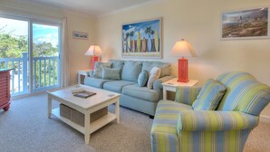 A living room with a sofa, striped armchair, and a coffee table. There are two lamps on side tables, a painting on the wall, and a view through a sliding door. - Enjoy beach walks and family fun in this 2BR, 2BA Oak Island condo. (Oak Island)
