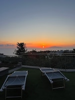 Una terraza en la azotea con dos tumbonas, vistas al atardecer sobre el mar y un paisaje urbano distante.