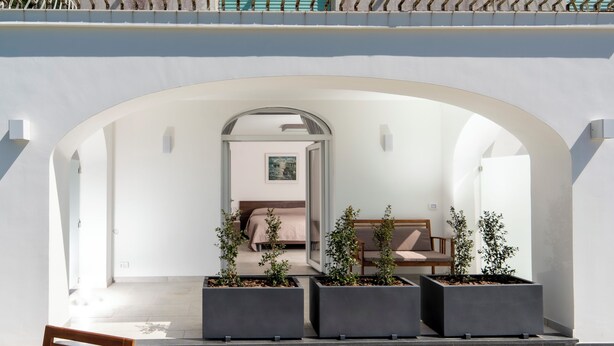 An outdoor seating area with a wooden chair and a bench, a white wall with an arched doorway, and three square planters with plants. - La Minucciola (Anacapri)