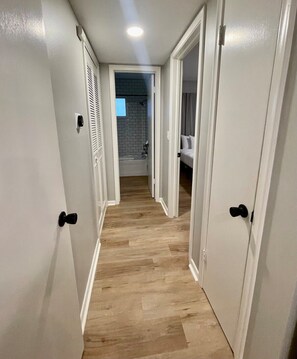 A narrow hallway with wooden flooring, white walls, and doors leading to a bathroom and a bedroom. - Stylish Midtown Apartment | Walk to Dining & UNMC (Omaha)