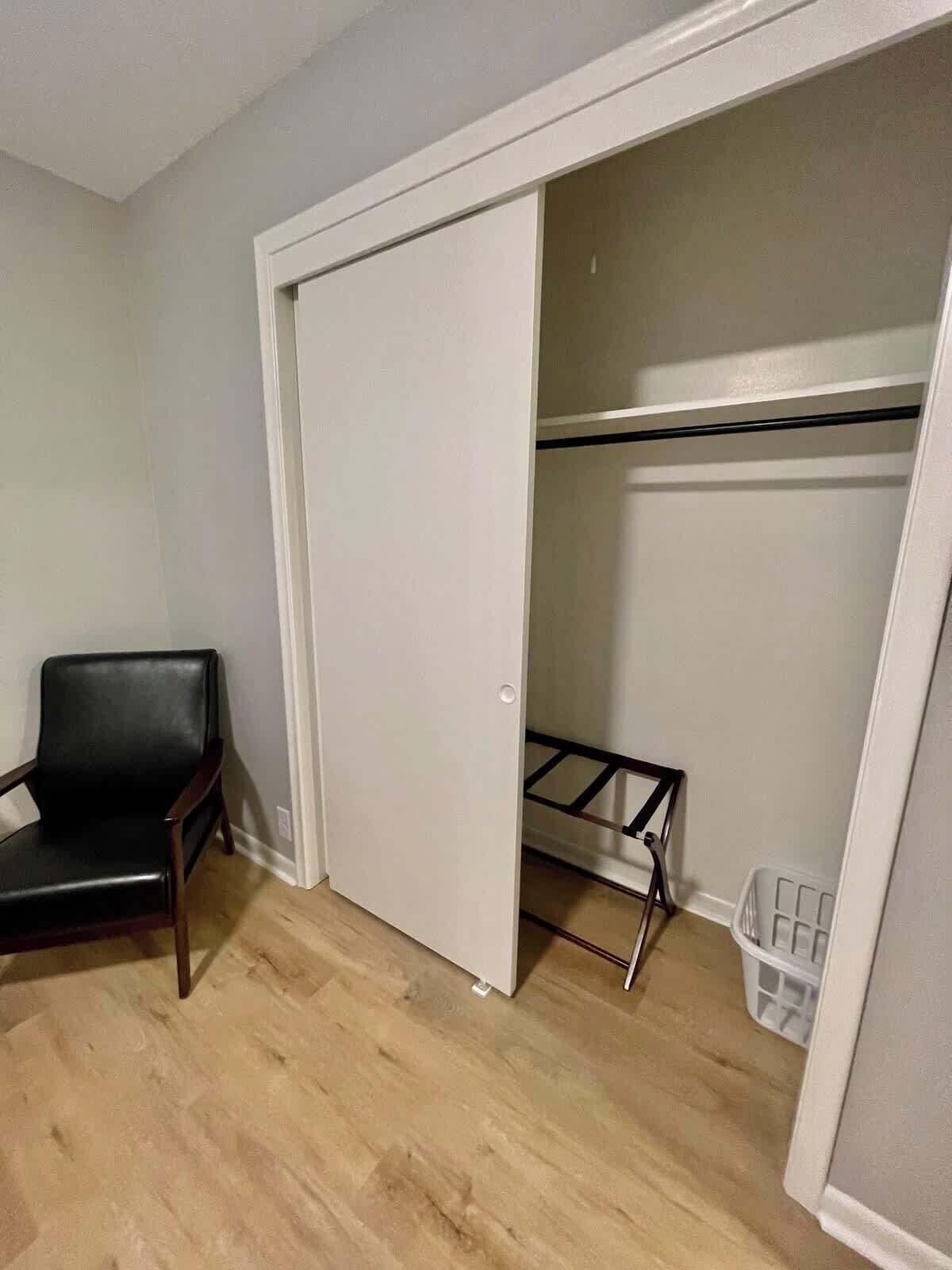 A room with a wooden floor, a black leather chair, a white closet with a black shelf, and a white laundry basket.