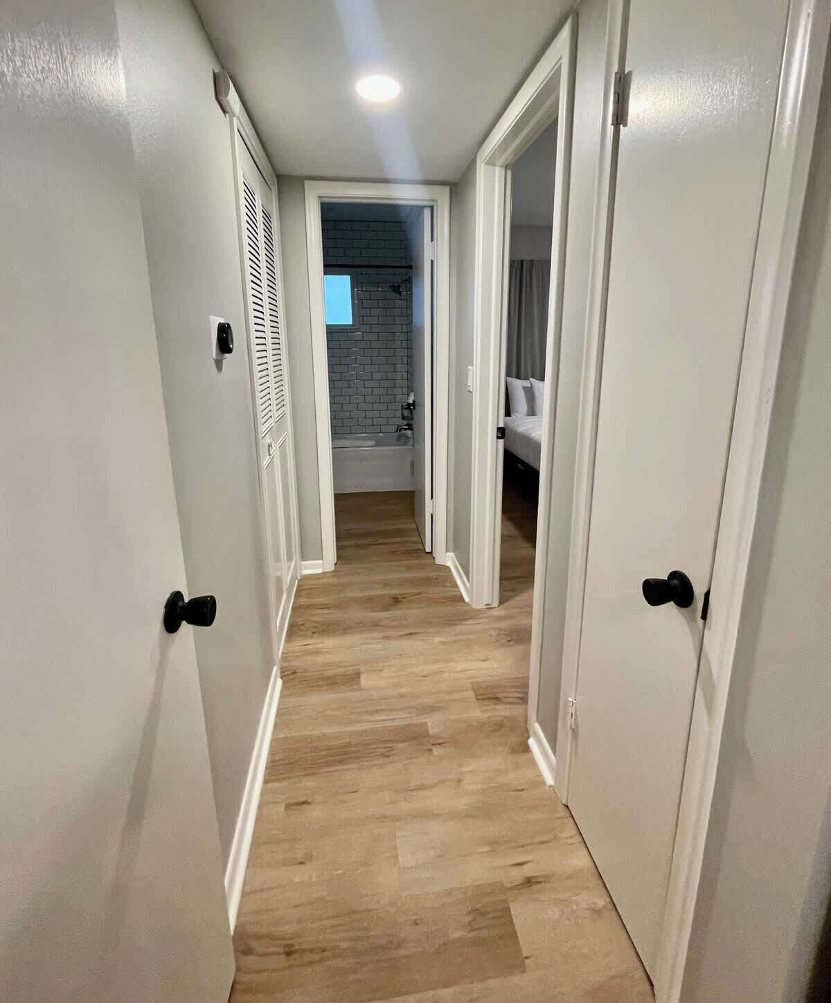 Un couloir étroit, doté d’un plancher en bois et de murs blancs, mène à une salle de bain équipée d’une baignoire et d’un espace douche.