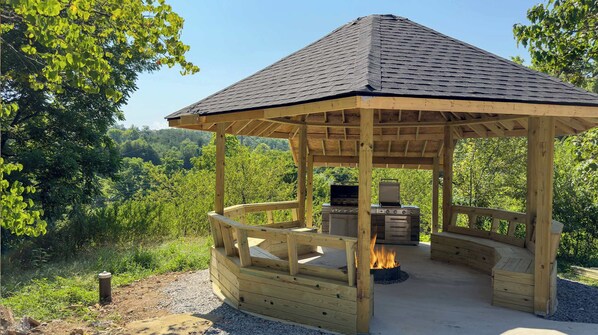 Un belvédère en bois doté d’une cheminée et d’un barbecue, entouré de verdure.