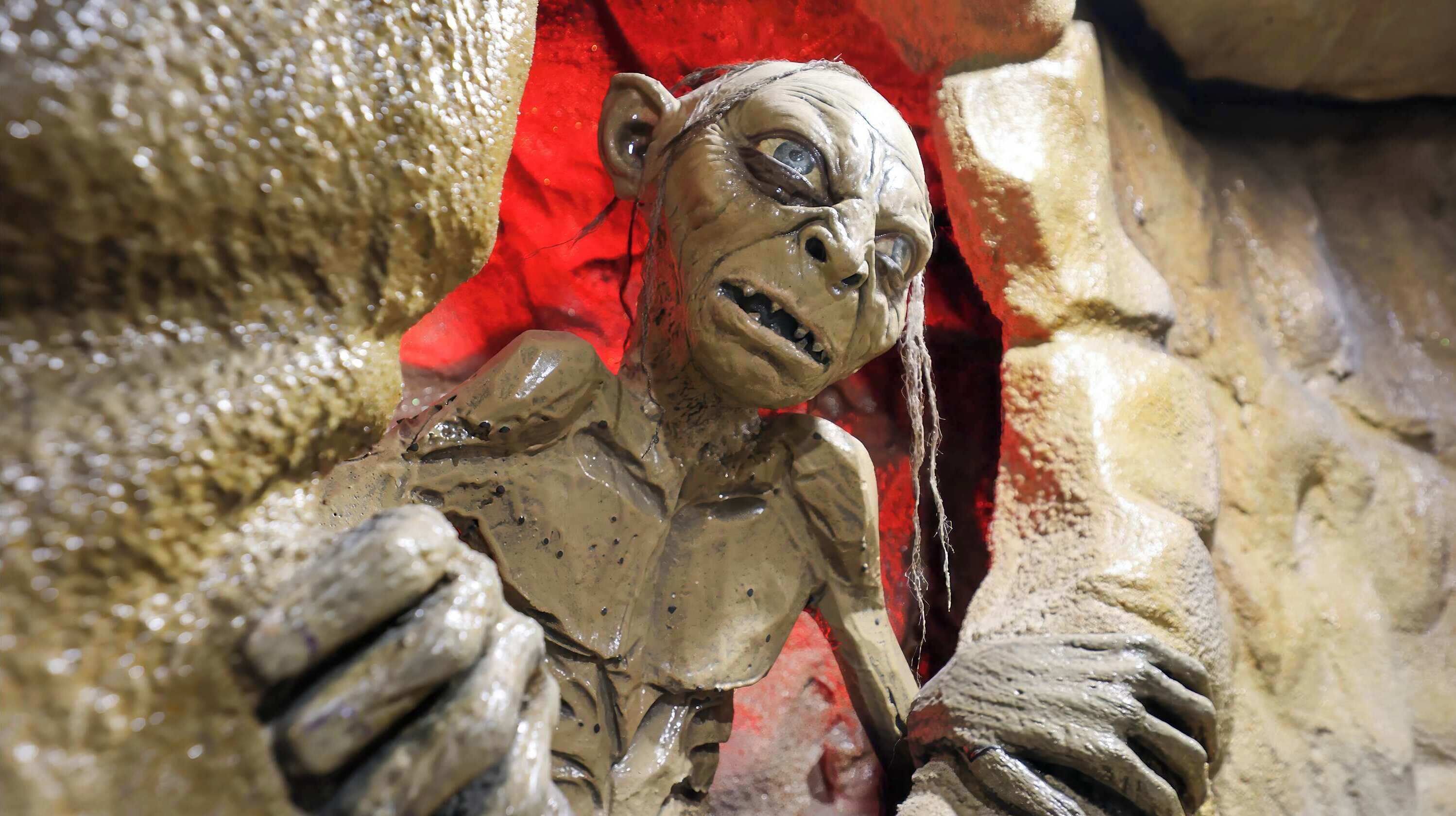 En skulptur af en menneskelignende figur med en detaljeret overflade, placeret i et klippefyldt landskab med rødt stof i baggrunden.