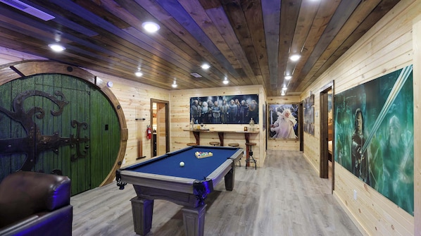 A pool table in a room with wooden walls and ceiling, a green door, and framed pictures on the walls.