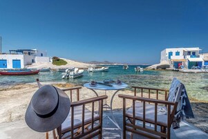 Vista al mar con una mesa y sillas, barcos y edificios blancos con detalles azules.