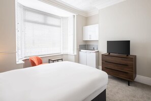 A bedroom with a bed, a TV on a wooden dresser, a small kitchenette, and a window with blinds.