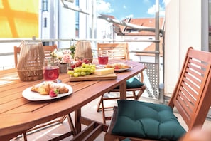 Et træbord med mad og drikke, fletkurve og grønne puder på en balkon med udsigt over byen.