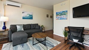 A living room with a sofa, ottoman, coffee table, TV, and a desk with a chair.