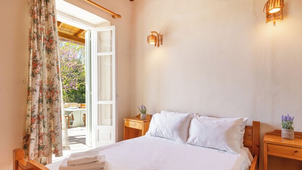 Une chambre à coucher avec un lit en bois, du linge de lit blanc, une table de chevet en bois avec une lampe, et une vue sur un jardin par une porte ouverte.