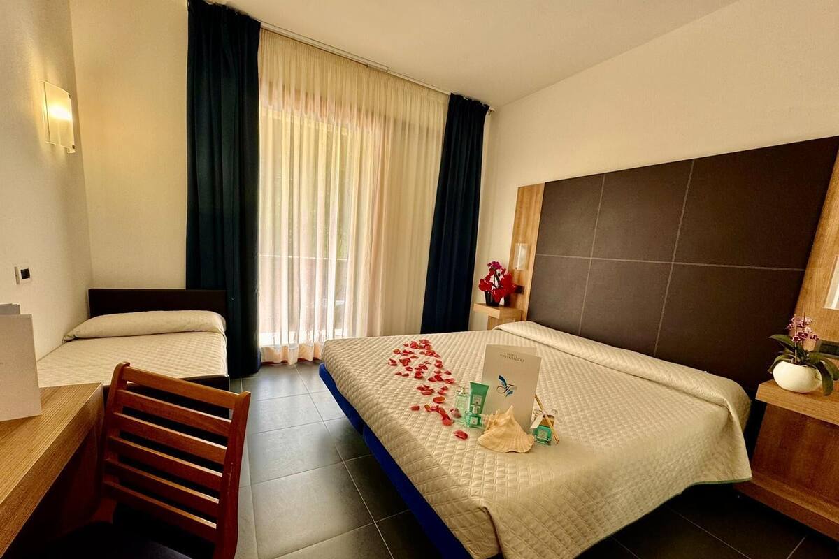 Een hotelkamer met twee bedden, een houten stoel, een nachtkastje met een bloevas en een groot raam met gordijnen.