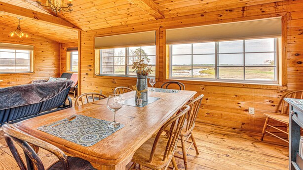 Interior de una cabaña de madera con una mesa de comedor, sillas, una araña y ventanas amplias que ofrecen vistas al exterior.
