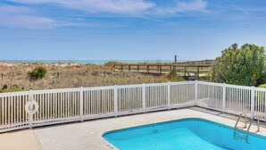 A swimming pool with a white fence, a lifebuoy, and a wooden deck leading to the beach.