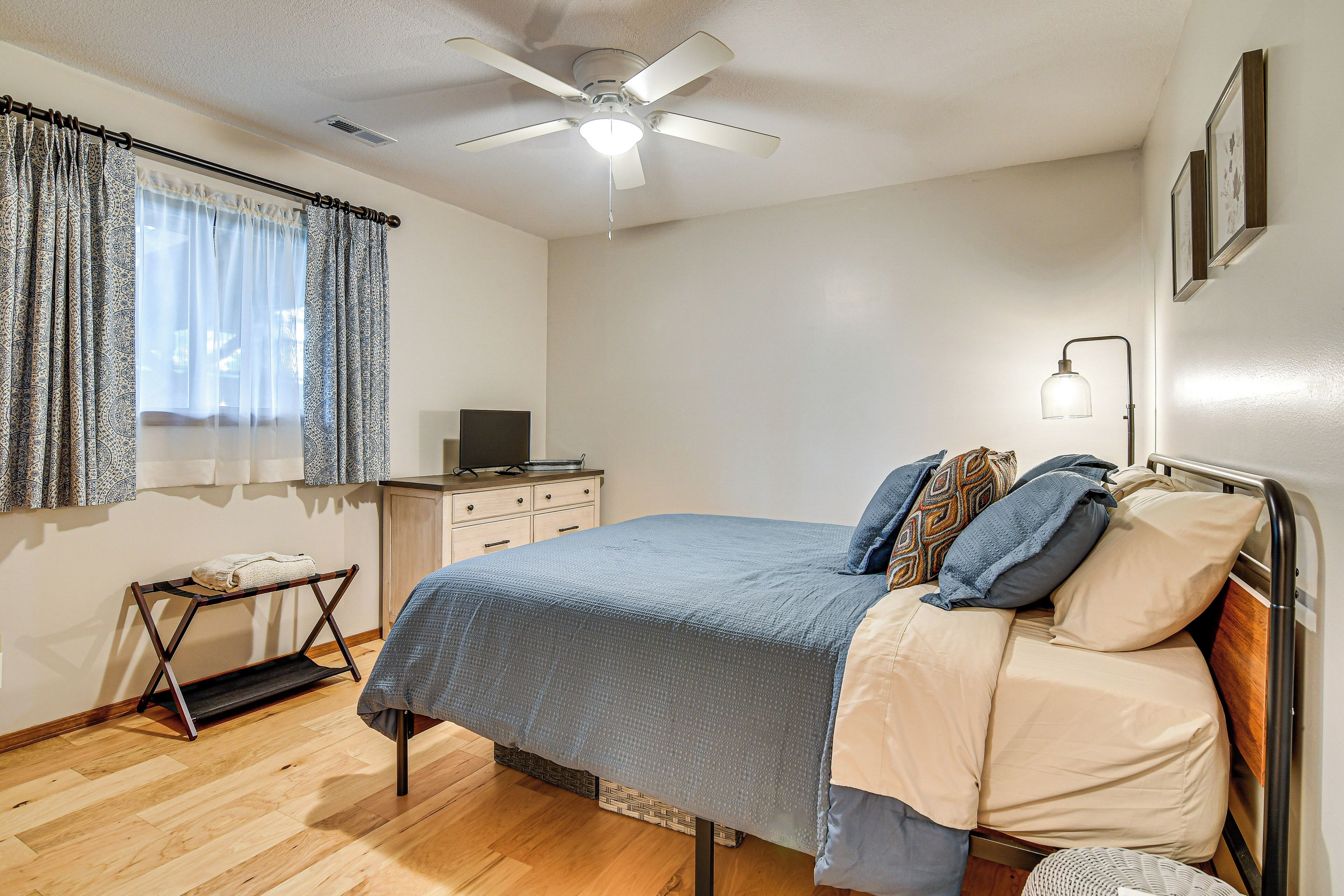 A bedroom with a bed, a ceiling fan, a bedside table with a lamp, a window with curtains, and a small desk with a computer.