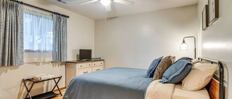 A bedroom with a bed, a ceiling fan, a bedside table with a lamp, a window with curtains, and a small desk with a computer.