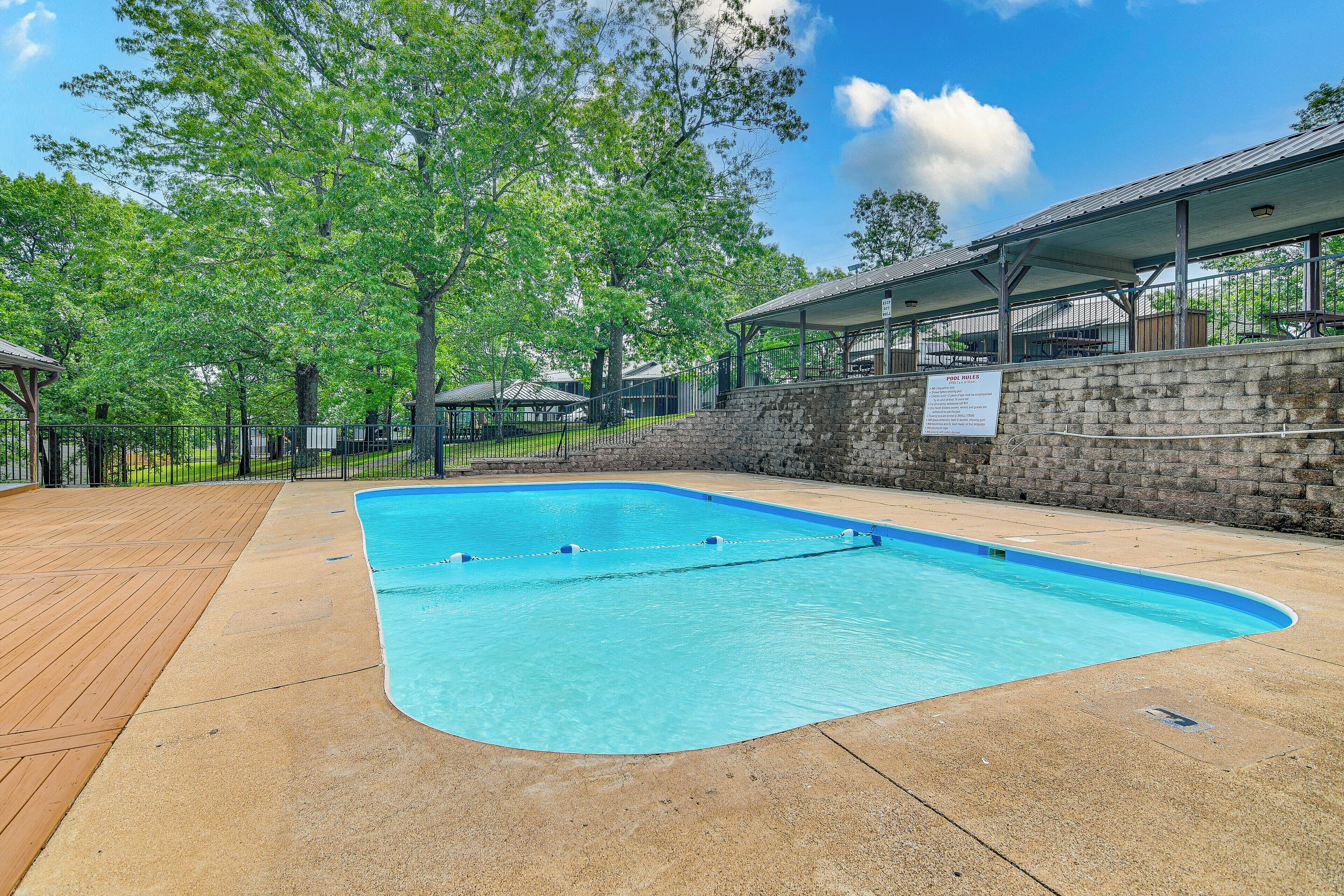 A swimming pool with a wooden deck and stone wall, surrounded by trees and covered seating areas.