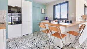 A modern kitchen with white cabinets, a built-in oven, and a window. There are three foldable chairs and a small table with cups and a kettle. - New house- 6 adults & 4 children - 350m beach (Noirmoutier-en-l´Ile)