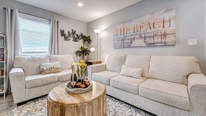 Un salón con dos sofás grises claros, una mesa de centro redonda de madera con una botella y flores, una pared con una obra de arte que dice 'THE BEACH', y una ventana con persianas.