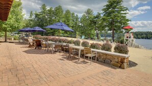 An outdoor patio with tables and chairs, umbrellas, and a stone wall. - Summer Village Family Cottage w/ Community Perks! (Westford)