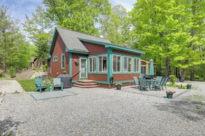 A small cabin with a covered patio, outdoor seating, and a gravel driveway.