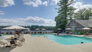 A pool area with lounge chairs and umbrellas, a building with a sign, and a forested background. - Summer Village Family Cottage w/ Community Perks! (Westford)