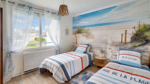 Une chambre avec une fresque murale représentant une plage, deux lits avec une literie assortie, une fenêtre avec des rideaux, un radiateur et une petite table de chevet avec une lampe.