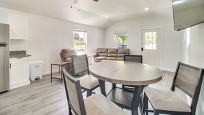 A spacious living area with a round dining table, comfortable seating, a ceiling fan, and large windows. - Walk to Lake: Cozy Grove Cabin w/ Fire Pit (Grove)